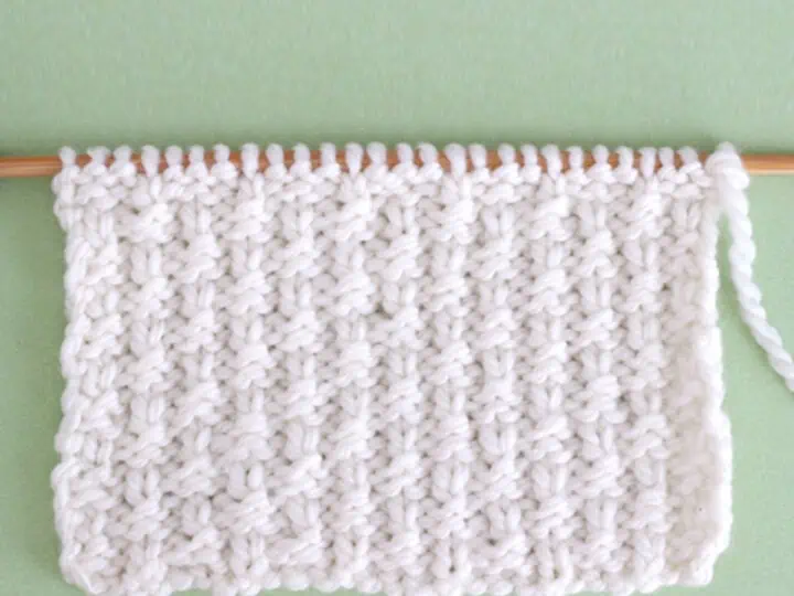 Little Raindrops Knit Stitch Pattern in white yarn on knitting needle atop a green background.