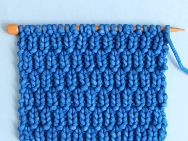 Long Raindrops Knit Stitch Pattern in bright blue yarn color on knitting needle atop a light blue background.