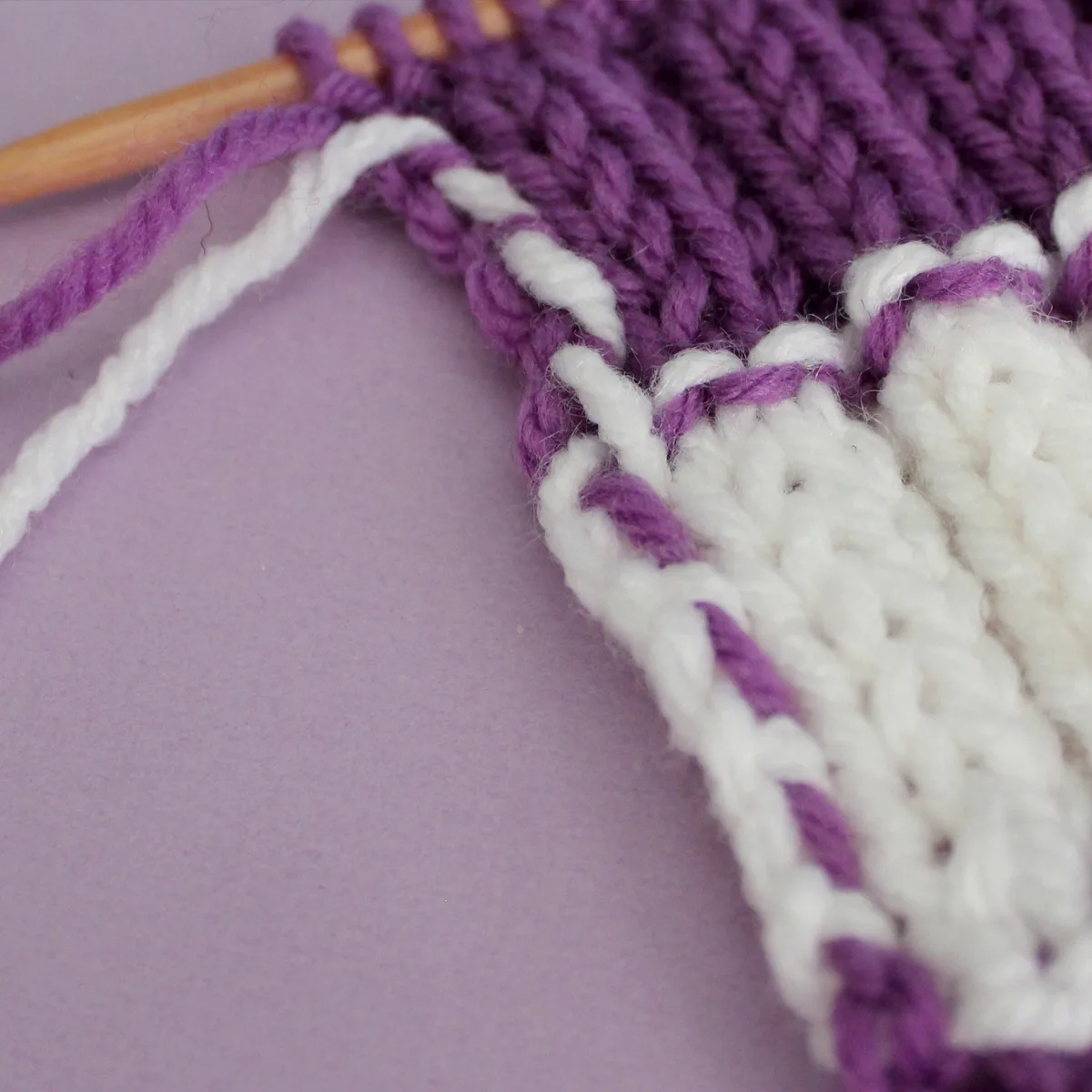 Yarn Carried up the side of a knitted swatch in stripes of purple and white colored yarn.