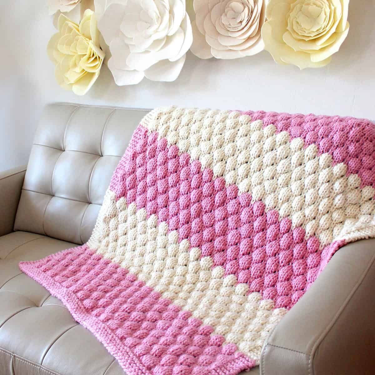 Knitted Bubble Stitch Blanket in pink and white yarn colors on a couch and felt flowers on the wall.