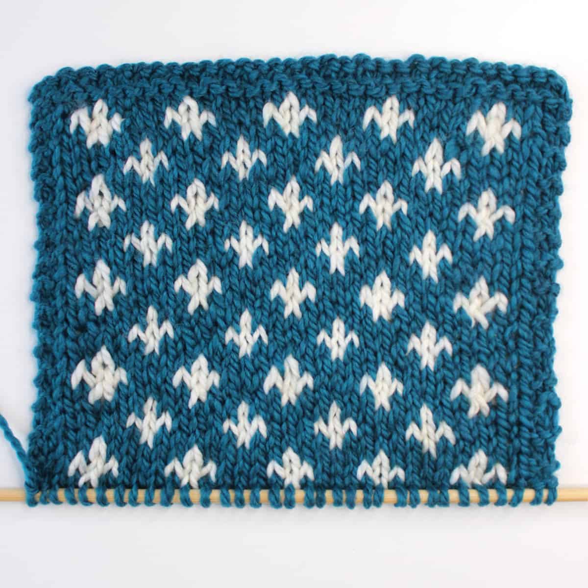 Fleur de Lys knit stitch pattern in blue and white color yarn on knitting needle.