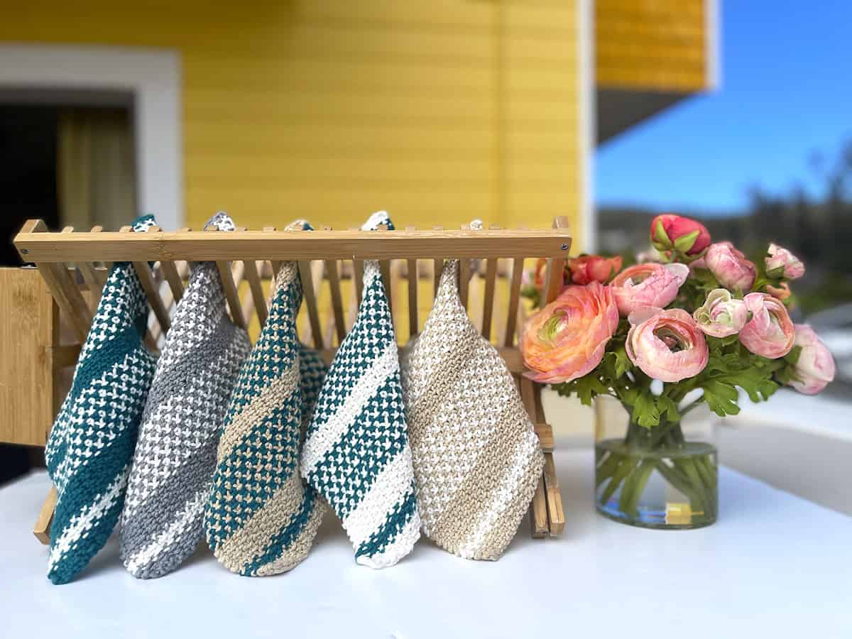 Five knitted dishcloths displayed on a dish rack with flowers.