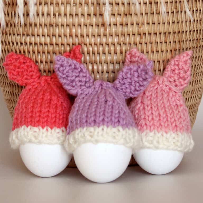 Knitted Bunny Egg Cozies in peach, purple, and pink yarn colors beside a wicker basket.