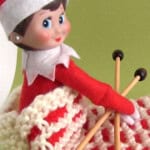 Elf on the shelf posing inside a knitted chimney sack holding needles.