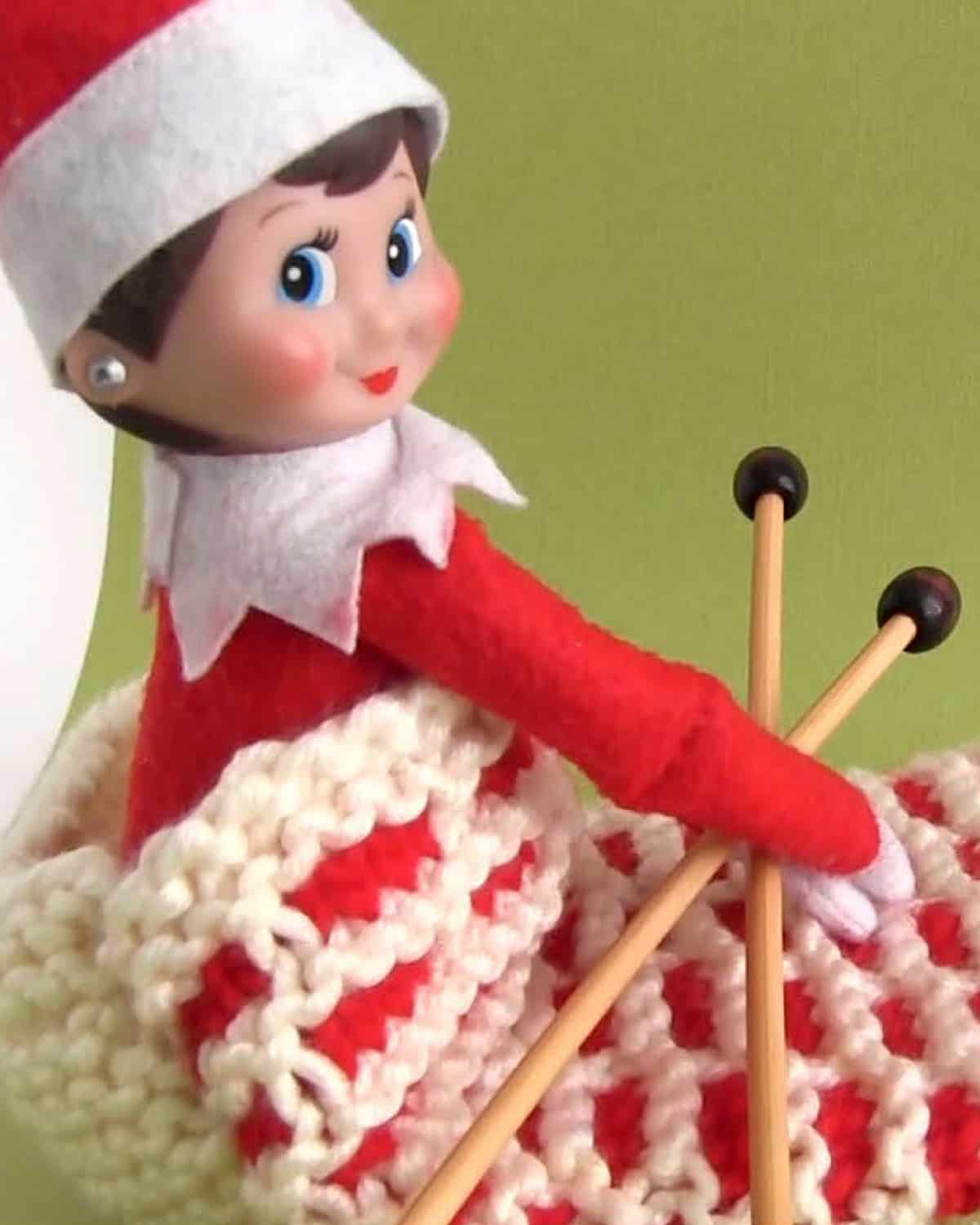 Elf on the shelf posing inside a knitted chimney sack holding needles.