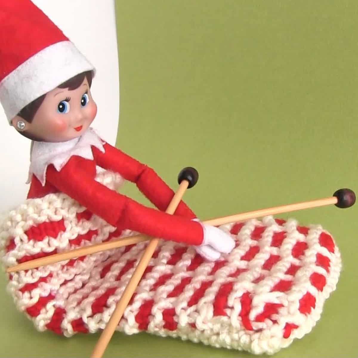 Elf on the shelf posing inside a knitted chimney sack holding needles.